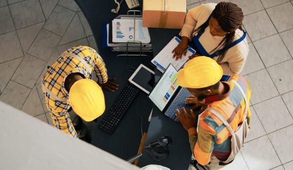 African american people looking at supplies list on pc, working with computer to check inventory and logistics. Employees organizing merchandise on shelves in warehouse space. Handheld shot.